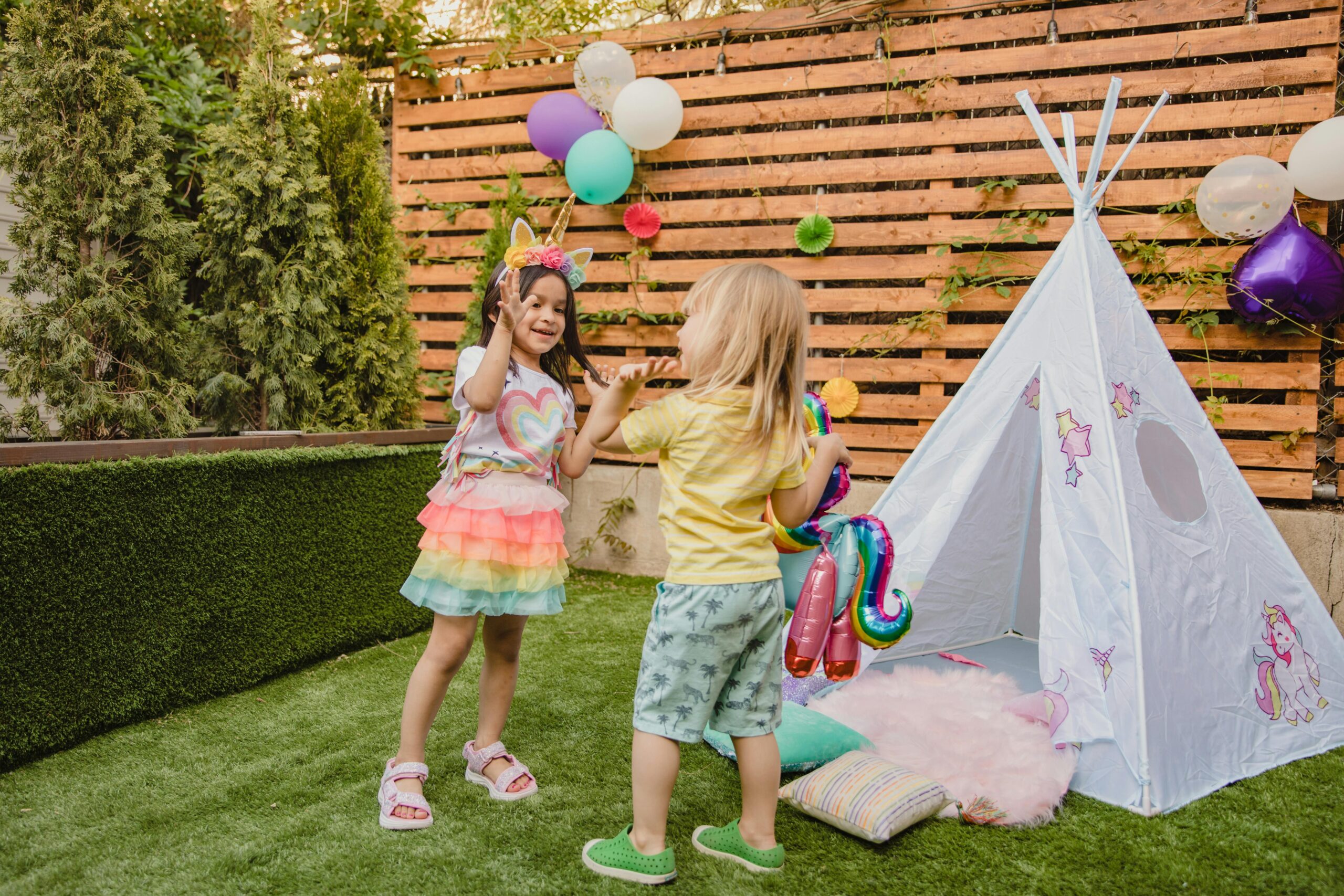 Two children play joyfully with balloons near a teepee tent, enjoying an outdoor party setting.
